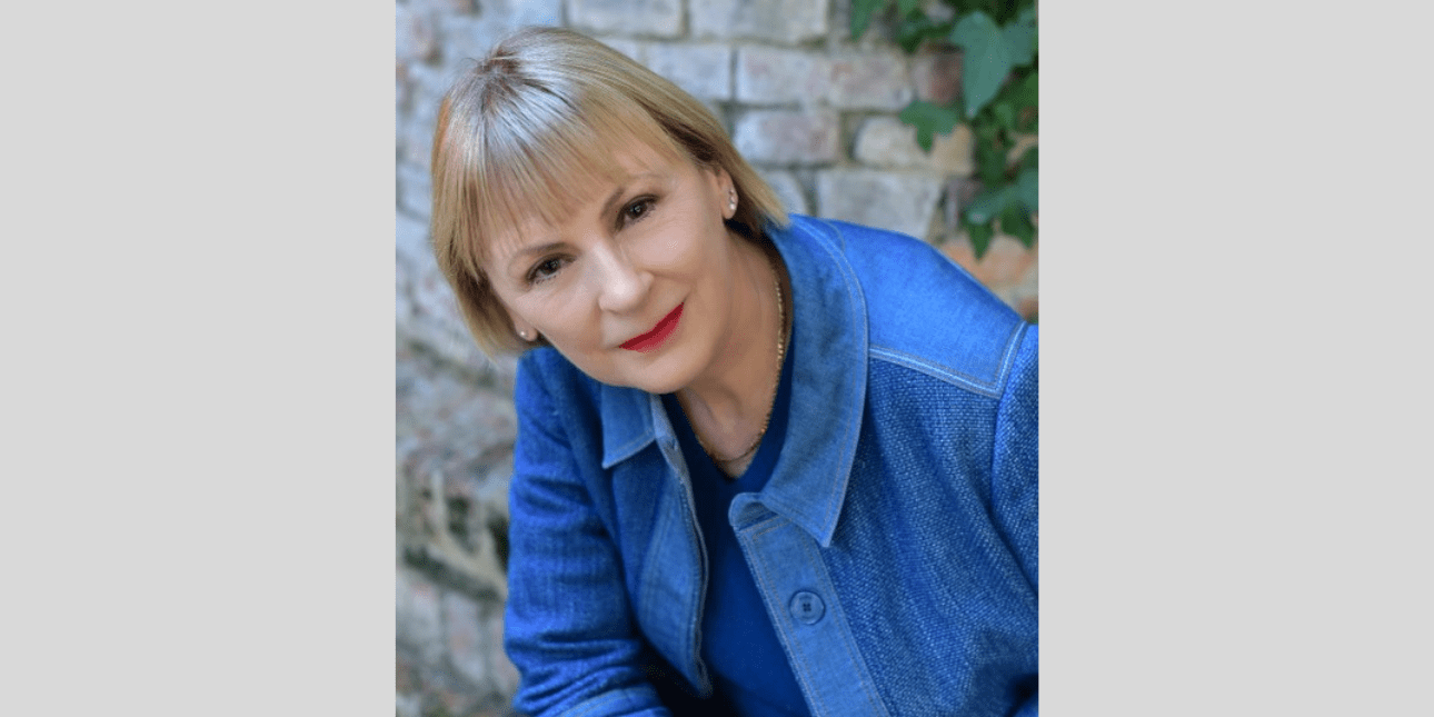 Portrait of Lindsay Nicholson, a white woman with short blonde hair wearing a blue shirt over a blue top sat with a brick wall and some foliage behind her